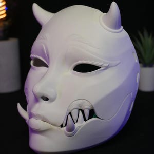 May include: A matte white mask featuring two small horns on the forehead, sculpted eyebrows, almond-shaped eye openings, and a lower jaw section with exposed sharp fangs and a single tusk protruding from the side of the mouth area.