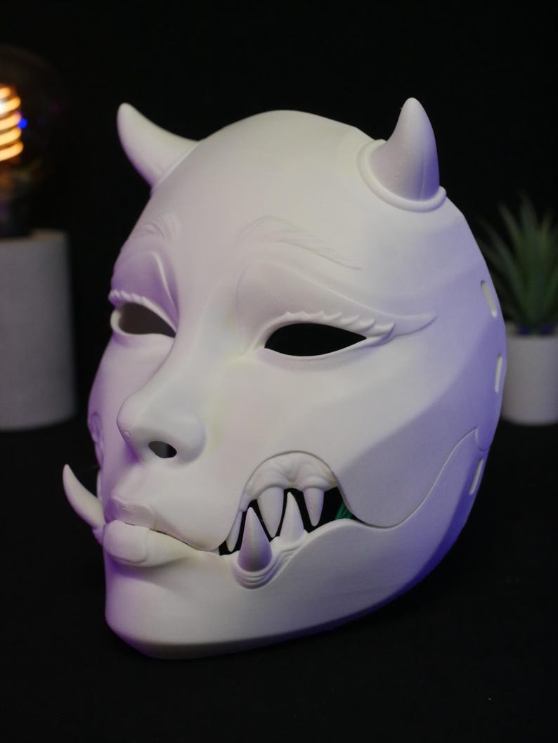 May include: A matte white mask featuring two small horns on the forehead, sculpted eyebrows, almond-shaped eye openings, and a lower jaw section with exposed sharp fangs and a single tusk protruding from the side of the mouth area.