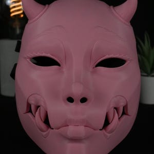 May include: A matte pink mask featuring two small horns at the top, almond-shaped eye openings, and a prominent mouth area with exposed fangs on both sides. The mask has a smooth, solid surface with sculpted facial features and a small chin detail.