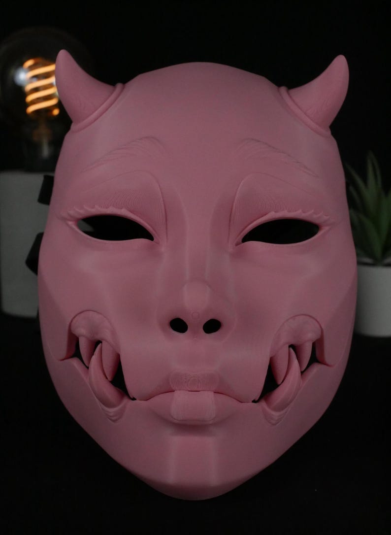 May include: A matte pink mask featuring two small horns at the top, almond-shaped eye openings, and a prominent mouth area with exposed fangs on both sides. The mask has a smooth, solid surface with sculpted facial features and a small chin detail.