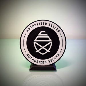 May include: A circular black and white sign featuring the text AUTHORIZED SELLER printed twice along the border. The center displays a white geometric logo consisting of stacked horizontal lines and a crossed shape, all mounted on a black base.