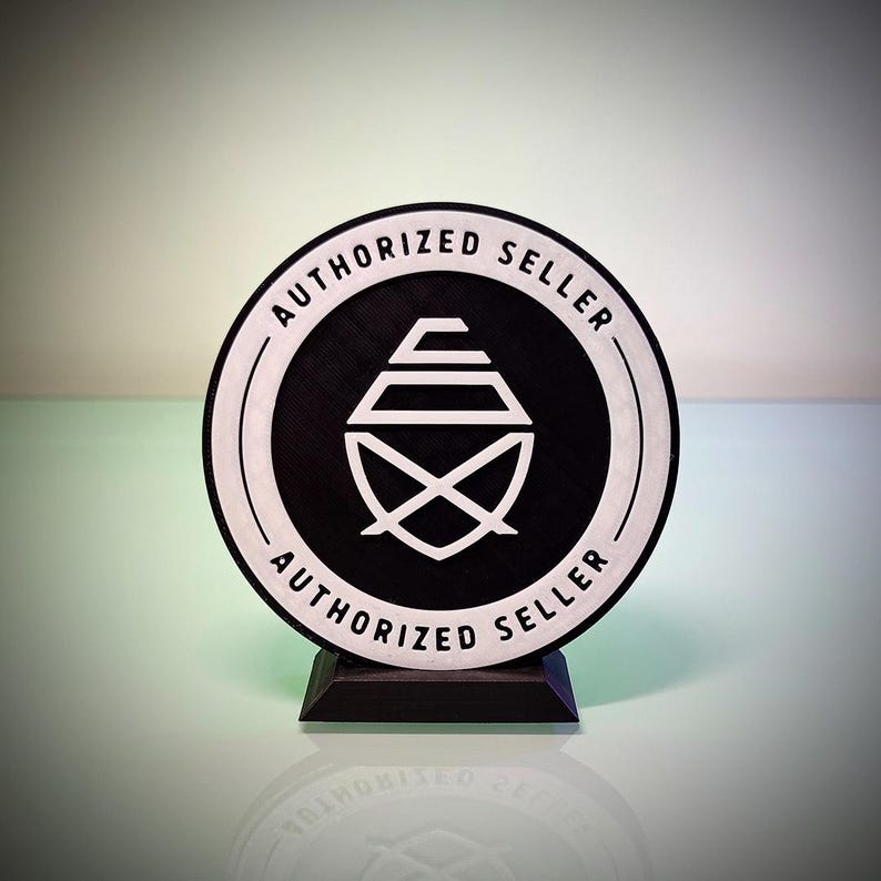 May include: A circular black and white sign featuring the text AUTHORIZED SELLER printed twice along the border. The center displays a white geometric logo consisting of stacked horizontal lines and a crossed shape, all mounted on a black base.