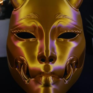 May include: A metallic gold mask featuring two small horns at the top, almond-shaped eye openings, and prominent fangs protruding from the sides of the mouth. The surface has a smooth, reflective finish with subtle purple highlights in the contours.