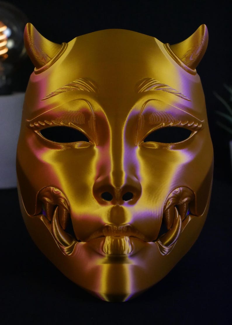 May include: A metallic gold mask featuring two small horns at the top, almond-shaped eye openings, and prominent fangs protruding from the sides of the mouth. The surface has a smooth, reflective finish with subtle purple highlights in the contours.