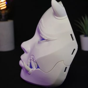 May include: A white, three-dimensional mask featuring a stylized face with a horn on the forehead and prominent fangs protruding from the mouth area. The mask has a smooth, matte finish with visible side vents and a blue light glowing from within.
