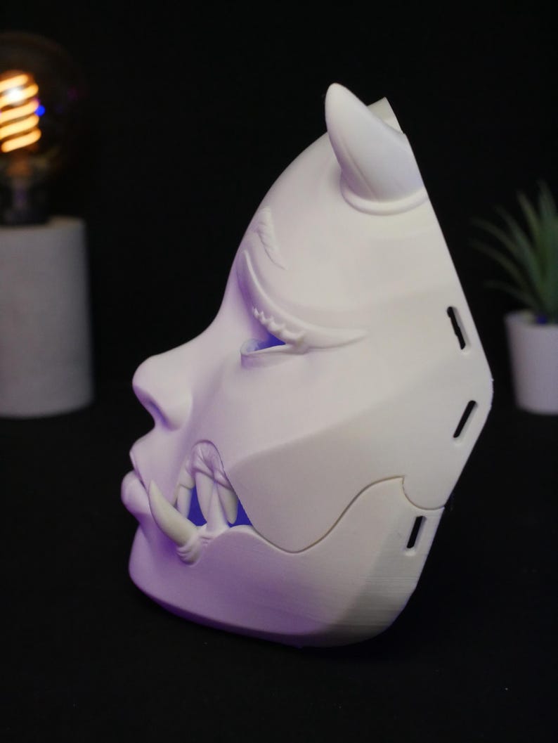 May include: A white, three-dimensional mask featuring a stylized face with a horn on the forehead and prominent fangs protruding from the mouth area. The mask has a smooth, matte finish with visible side vents and a blue light glowing from within.