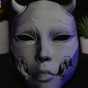 May include: A light gray, 3D-printed mask featuring a stylized human face with two small horns on the forehead, almond-shaped eyes, and prominent fangs protruding from the corners of the mouth. The surface shows fine horizontal layer lines throughout.