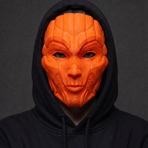 Peut inclure: Un masque intégral orange vif présentant des détails sculptés complexes, superposés et géométriques. Le masque est doté d'ouvertures profondes pour les yeux et d'une surface structurée et segmentée, porté avec un sweat à capuche noir uni.