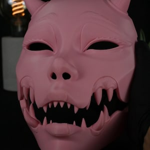 May include: A solid pink mask featuring a stylized creature face with two pointed horns on the forehead, almond-shaped eye openings, and a wide, open mouth revealing multiple sharp, protruding teeth along the upper and lower jaw lines.