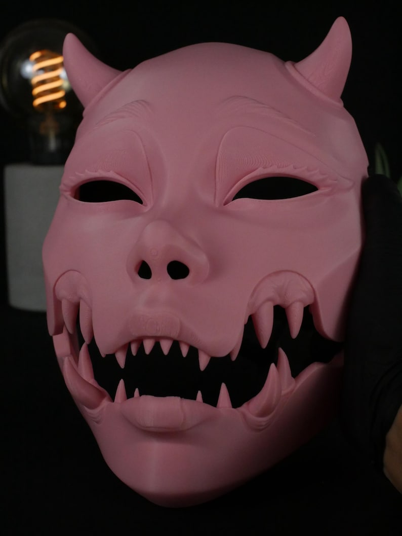 May include: A solid pink mask featuring a stylized creature face with two pointed horns on the forehead, almond-shaped eye openings, and a wide, open mouth revealing multiple sharp, protruding teeth along the upper and lower jaw lines.
