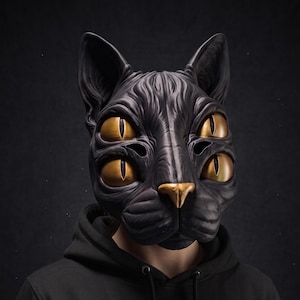 Puede incluir: Una máscara de gato negro con cuatro ojos dorados de pupilas verticales y una nariz dorada. La máscara presenta una textura de pelaje detallada y orejas puntiagudas, usada sobre una sudadera con capucha negra lisa. El diseño es simétrico.