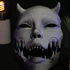 May include: A matte gray, 3D-printed mask featuring a stylized creature design with two small horns on the forehead, almond-shaped eyes, and a wide, open mouth filled with sharp, pointed teeth. The surface shows fine horizontal layer lines.