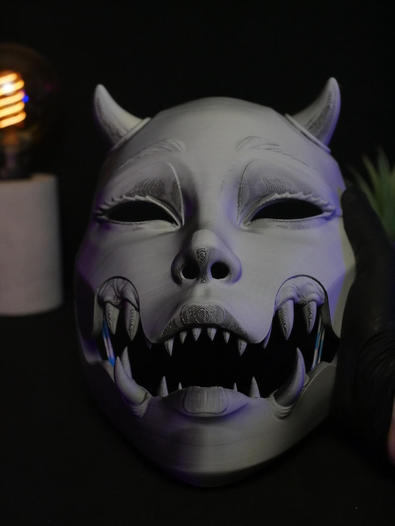 May include: A matte gray, 3D-printed mask featuring a stylized creature design with two small horns on the forehead, almond-shaped eyes, and a wide, open mouth filled with sharp, pointed teeth. The surface shows fine horizontal layer lines.