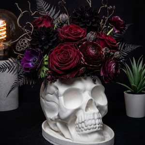 Può includere: Un vaso a forma di teschio bianco poggiato su una base ovale abbinata, riempito con una composizione floreale scura composta da rose rosso scuro, dalie viola scuro, anemoni, baccelli secchi e foglie di felce nere.