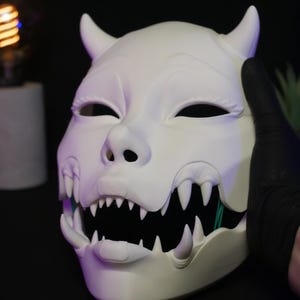 May include: A matte white, full-face mask featuring two prominent horns at the top, almond-shaped eye openings, and a wide, open mouth revealing a set of sharp, pointed teeth. The mask has a smooth, sculpted surface with distinct facial features.