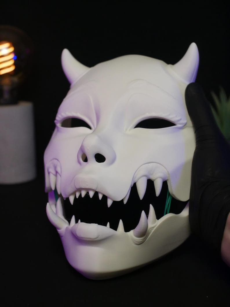 May include: A matte white, full-face mask featuring two prominent horns at the top, almond-shaped eye openings, and a wide, open mouth revealing a set of sharp, pointed teeth. The mask has a smooth, sculpted surface with distinct facial features.