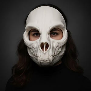 Può includere: Una maschera a teschio bianca con una struttura ossea dettagliata, tra cui orbite, cavità nasale e denti. La maschera è indossata da una persona con capelli scuri e un top nero, su uno sfondo grigio scuro. La maschera è un accessorio di costume.