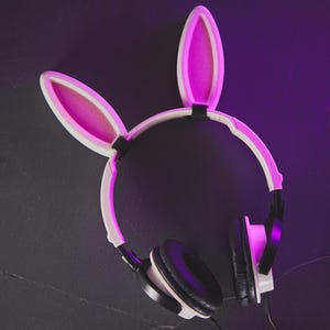 May include: White headphones with black earcups and a bunny ear headband. The headband and ear interiors are pink. The headphones are on a dark surface with a purple hue.