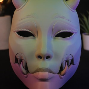 May include: A full-face mask featuring a gradient of pastel green, blue, and purple hues. The mask displays two small horns at the top, almond-shaped eye openings, and a lower section revealing sharp fangs and a small tongue protruding from the mouth.