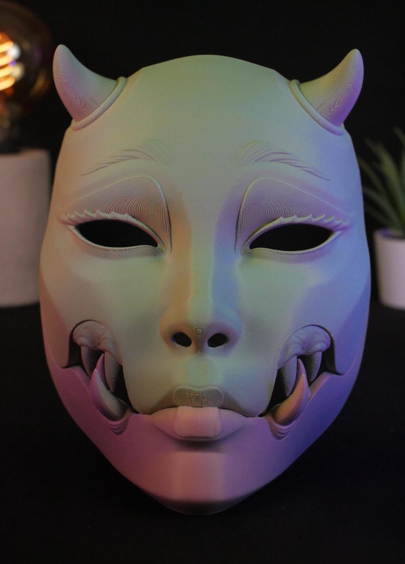 May include: A full-face mask featuring a gradient of pastel green, blue, and purple hues. The mask displays two small horns at the top, almond-shaped eye openings, and a lower section revealing sharp fangs and a small tongue protruding from the mouth.