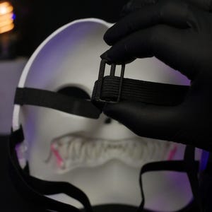 May include: A white mask featuring a jagged, toothy mouth design with pink accents. The mask includes black elastic straps with a black plastic adjustable buckle for securing the fit, shown from the back side against a dark background.
