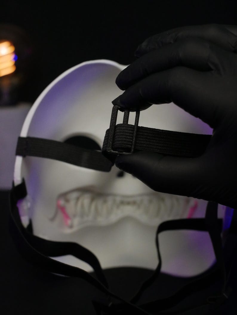 May include: A white mask featuring a jagged, toothy mouth design with pink accents. The mask includes black elastic straps with a black plastic adjustable buckle for securing the fit, shown from the back side against a dark background.