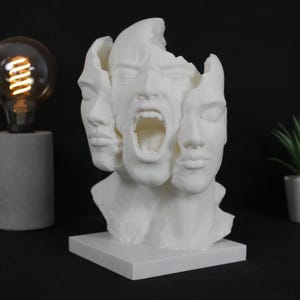 Könnte beinhalten: Eine weiße Skulptur eines Gesichts mit drei sichtbaren Ausdrücken: ruhig, neutral und schreiend. Die Skulptur steht auf einem quadratischen Sockel vor einem dunklen Hintergrund. Ein einzigartiges Kunstwerk.