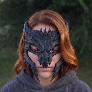 Op de afbeelding: Een matzwart, op een draak geïnspireerd masker met ingewikkelde, getextureerde schubpatronen, scherpe, hoornachtige uitsteeksels langs het voorhoofd en puntige, slagtandachtige structuren die over de jukbeenderen naar beneden lopen en het gezicht bedekken.