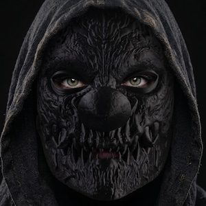 Könnte beinhalten: Eine schwarze, texturierte Maske mit einer runden Nase und spitzen Zähnen, die den unteren Gesichtsbereich bedeckt und unter einer dunklen Kapuze getragen wird. Die Maske weist tiefe, komplexe Rillen auf und zeigt hellgrüne Augen.