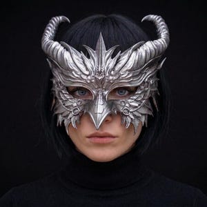 May include: A silver-coloured mask featuring intricate, wing-like filigree patterns and two prominent, curved horns extending from the top. The mask covers the upper portion of the face, including the eyes and nose, with a sharp, pointed central bridge.