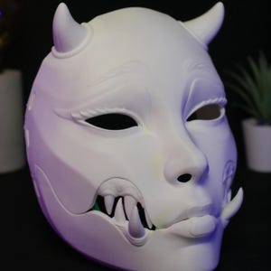 May include: A matte white, full-face mask featuring two small horns on the forehead, almond-shaped eye openings, and a protruding lower jaw with sharp, visible fangs. The mask has a smooth, sculpted surface with detailed facial features.