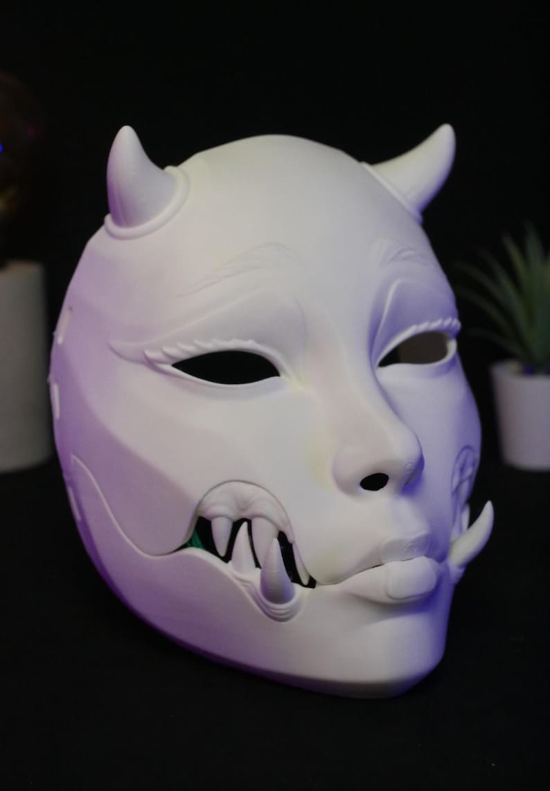 May include: A matte white, full-face mask featuring two small horns on the forehead, almond-shaped eye openings, and a protruding lower jaw with sharp, visible fangs. The mask has a smooth, sculpted surface with detailed facial features.