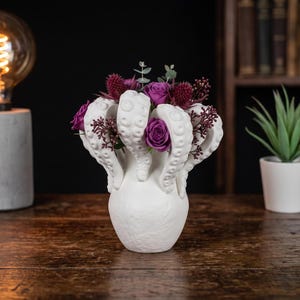 Peut inclure: Un vase en céramique blanche orné de tentacules de pieuvre sculptés sur le bord, contenant un arrangement floral composé de roses violettes, de fleurs épineuses rouge foncé et de petites feuilles d'eucalyptus vertes.