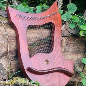 May include: A reddish-brown lyre with silver tuning pegs and strings. The instrument has a curved shape with a sound hole and a dark brown accent piece. The lyre is set against a backdrop of wood and green foliage.