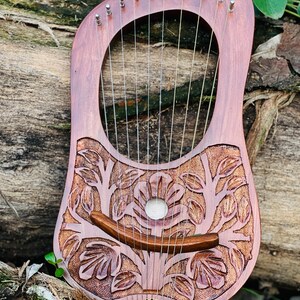 K&ouml;nnte beinhalten: Eine Holzbogenharfe mit 10 Saiten, mit einem geschnitzten Blumendesign. Das Instrument hat eine rotbraune Farbe, mit einem gebogenen Steg und Stimmwirbeln oben. Die Leier liegt auf einer nat&uuml;rlichen Holzoberfl&auml;che.