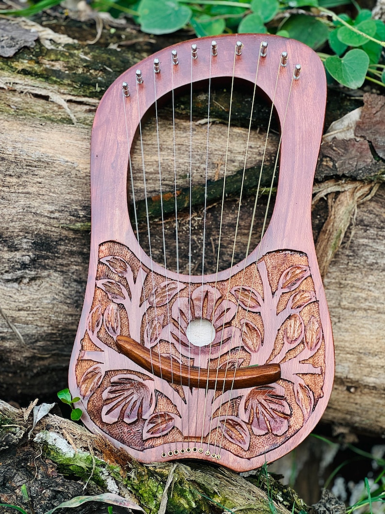 K&ouml;nnte beinhalten: Eine Holzbogenharfe mit 10 Saiten, mit einem geschnitzten Blumendesign. Das Instrument hat eine rotbraune Farbe, mit einem gebogenen Steg und Stimmwirbeln oben. Die Leier liegt auf einer nat&uuml;rlichen Holzoberfl&auml;che.