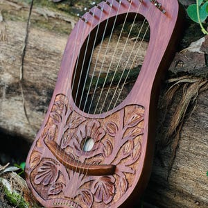 K&ouml;nnte beinhalten: Eine handgefertigte Holzleier mit einem geschnitzten Blumendesign. Das Instrument hat eine satte, rotbraune Oberfl&auml;che und zahlreiche Saiten. Sie liegt auf einem verwitterten Baumstamm, mit gr&uuml;nem Laub im Hintergrund. Das Schallloch der Leier ist sichtbar.