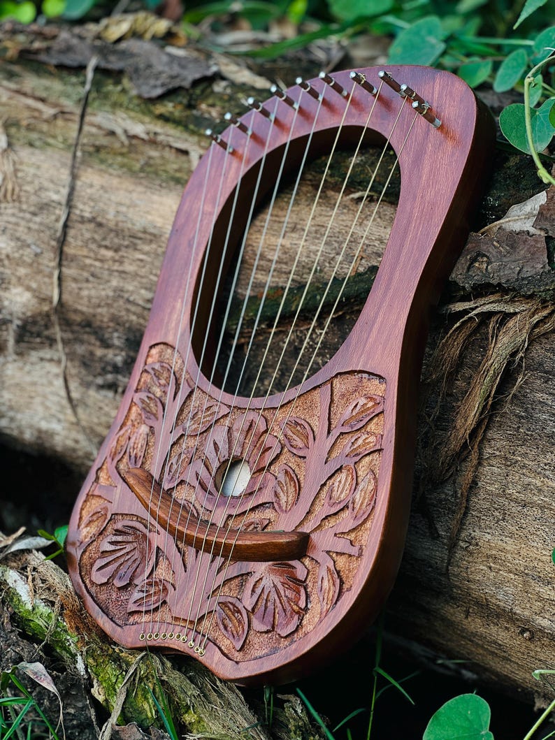 K&ouml;nnte beinhalten: Eine handgefertigte Holzleier mit einem geschnitzten Blumendesign. Das Instrument hat eine satte, rotbraune Oberfl&auml;che und zahlreiche Saiten. Sie liegt auf einem verwitterten Baumstamm, mit gr&uuml;nem Laub im Hintergrund. Das Schallloch der Leier ist sichtbar.