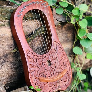 K&ouml;nnte beinhalten: Eine handgefertigte, rotbraune Lyra mit aufwendigen Blumenschnitzereien. Das Instrument verf&uuml;gt &uuml;ber mehrere Saiten und einen gebogenen Holzsteg. Die Lyra ist vor einem nat&uuml;rlichen Hintergrund aus Holz und Gr&uuml;n platziert, was ihre organische &Auml;sthetik hervorhebt.