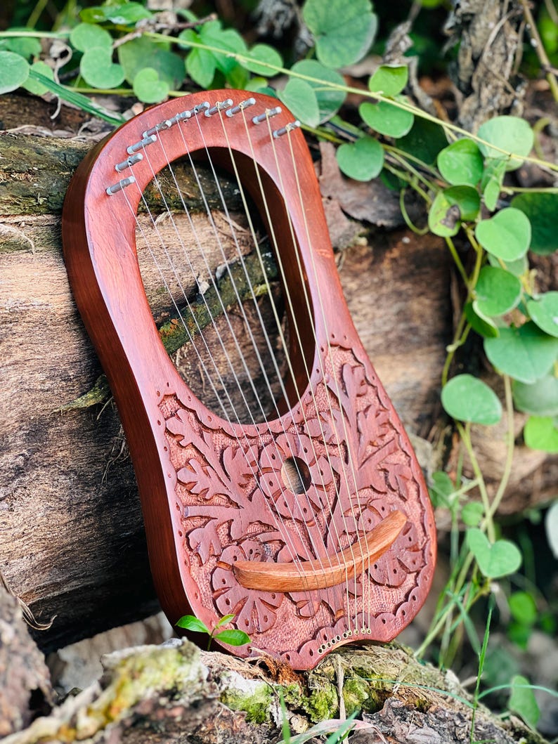 K&ouml;nnte beinhalten: Eine handgefertigte, rotbraune Lyra mit aufwendigen Blumenschnitzereien. Das Instrument verf&uuml;gt &uuml;ber mehrere Saiten und einen gebogenen Holzsteg. Die Lyra ist vor einem nat&uuml;rlichen Hintergrund aus Holz und Gr&uuml;n platziert, was ihre organische &Auml;sthetik hervorhebt.