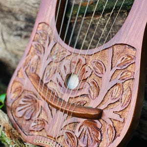 K&ouml;nnte beinhalten: Eine handgefertigte Holzleier mit aufwendigen Blumenschnitzereien. Das Instrument hat eine warme braune Oberfl&auml;che, mehrere Saiten und ein Schallloch. Das Design umfasst detaillierte Blatt- und Astmuster, die die Handwerkskunst des Instruments zeigen.