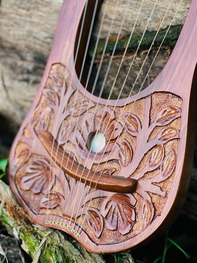 K&ouml;nnte beinhalten: Eine handgefertigte Holzleier mit aufwendigen Blumenschnitzereien. Das Instrument hat eine warme braune Oberfl&auml;che, mehrere Saiten und ein Schallloch. Das Design umfasst detaillierte Blatt- und Astmuster, die die Handwerkskunst des Instruments zeigen.