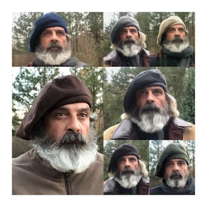 Puede incluir: Una colección de boinas en varios colores, incluyendo azul marino, gris, marrón y verde oliva. Las boinas son usadas por una persona con una larga barba blanca. El fondo presenta árboles y luz natural, creando una estética rústica.