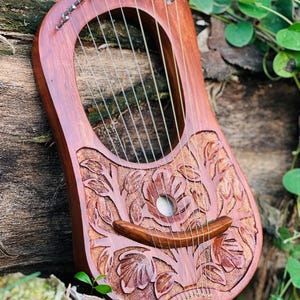 K&ouml;nnte beinhalten: Eine Holzleier mit einem geschnitzten Blumendesign. Das Instrument hat eine rotbraune Oberfl&auml;che, mit Saiten, die &uuml;ber dem Schallloch gespannt sind. Die Leier liegt auf einem Holzst&uuml;ck, mit gr&uuml;nem Laub im Hintergrund.