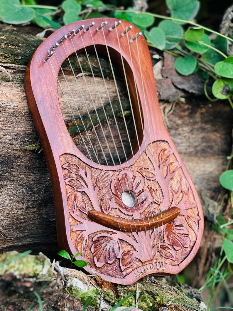 K&ouml;nnte beinhalten: Eine Holzleier mit einem geschnitzten Blumendesign. Das Instrument hat eine rotbraune Oberfl&auml;che, mit Saiten, die &uuml;ber dem Schallloch gespannt sind. Die Leier liegt auf einem Holzst&uuml;ck, mit gr&uuml;nem Laub im Hintergrund.