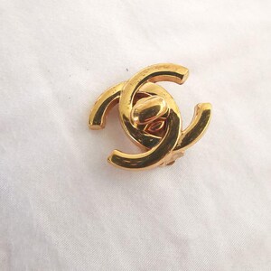 Puede incluir: Un amuleto de logotipo CC entrelazado de color dorado. El amuleto est&aacute; hecho de metal y tiene un acabado pulido.
