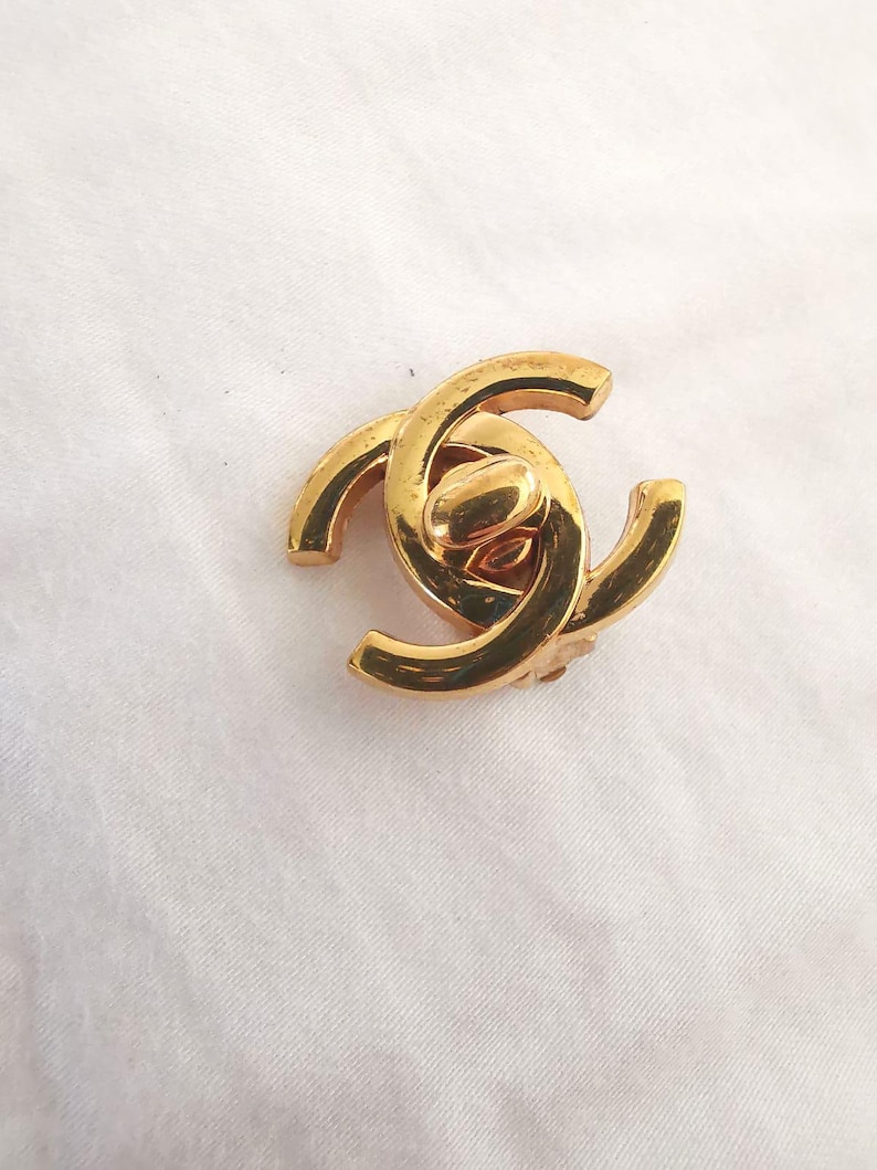 Puede incluir: Un amuleto de logotipo CC entrelazado de color dorado. El amuleto est&aacute; hecho de metal y tiene un acabado pulido.