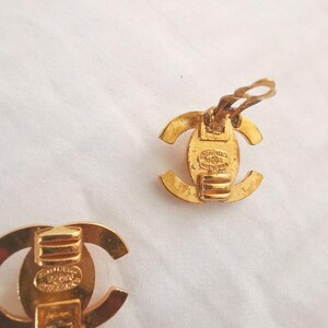 Puede incluir: Un par de pendientes Chanel de tono dorado con un logotipo CC. Los pendientes son de estilo clip y tienen un acabado texturizado.