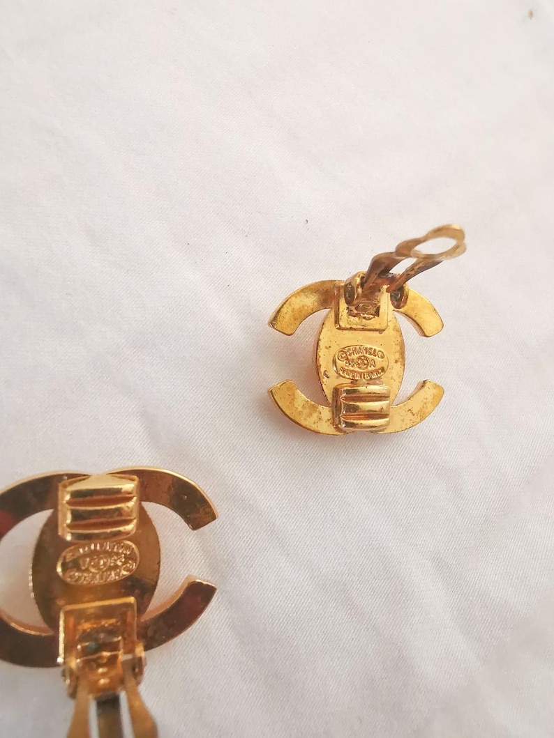 Puede incluir: Un par de pendientes Chanel de tono dorado con un logotipo CC. Los pendientes son de estilo clip y tienen un acabado texturizado.