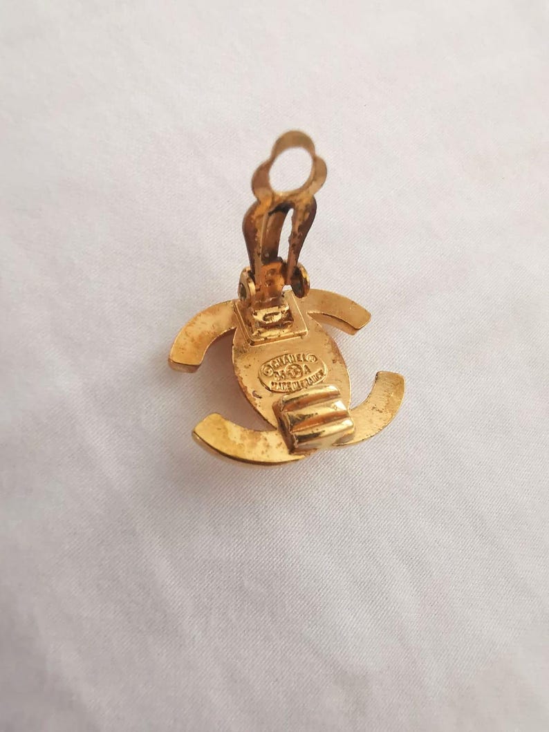 Puede incluir: Pendientes de clip Chanel de color dorado con el logotipo de Chanel en la parte posterior. Los pendientes est&aacute;n hechos de metal y tienen un dise&ntilde;o cl&aacute;sico.